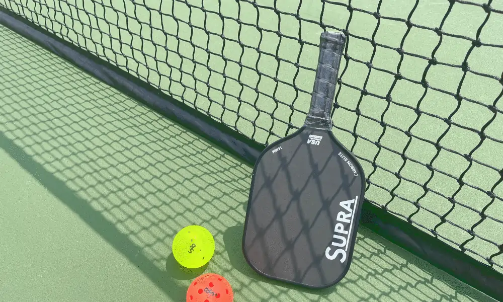 Pickleball Schlaeger mit Schutzhuelle und gelben und orangem Pickleball auf Pickleplatzgrund und schwarzem Pickleballnetz